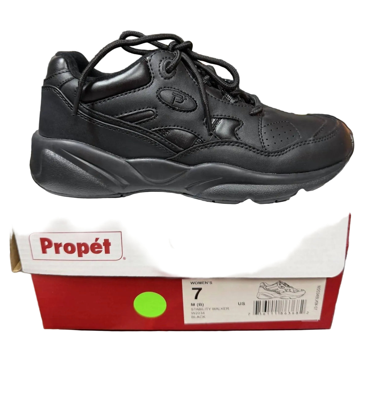 A black leather Pépét sneaker with black laces, resting on top of its white and red Propét shoebox, set on a desk with a keyboard and office items in the background.