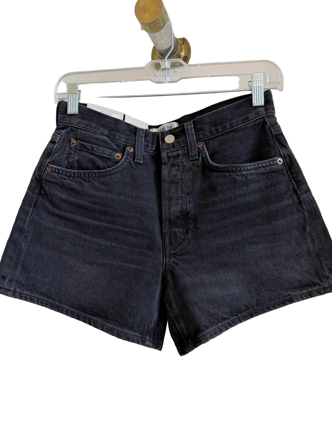 A pair of dark washed denim shorts hanging from a clear plastic hanger against a plain off-white wall.